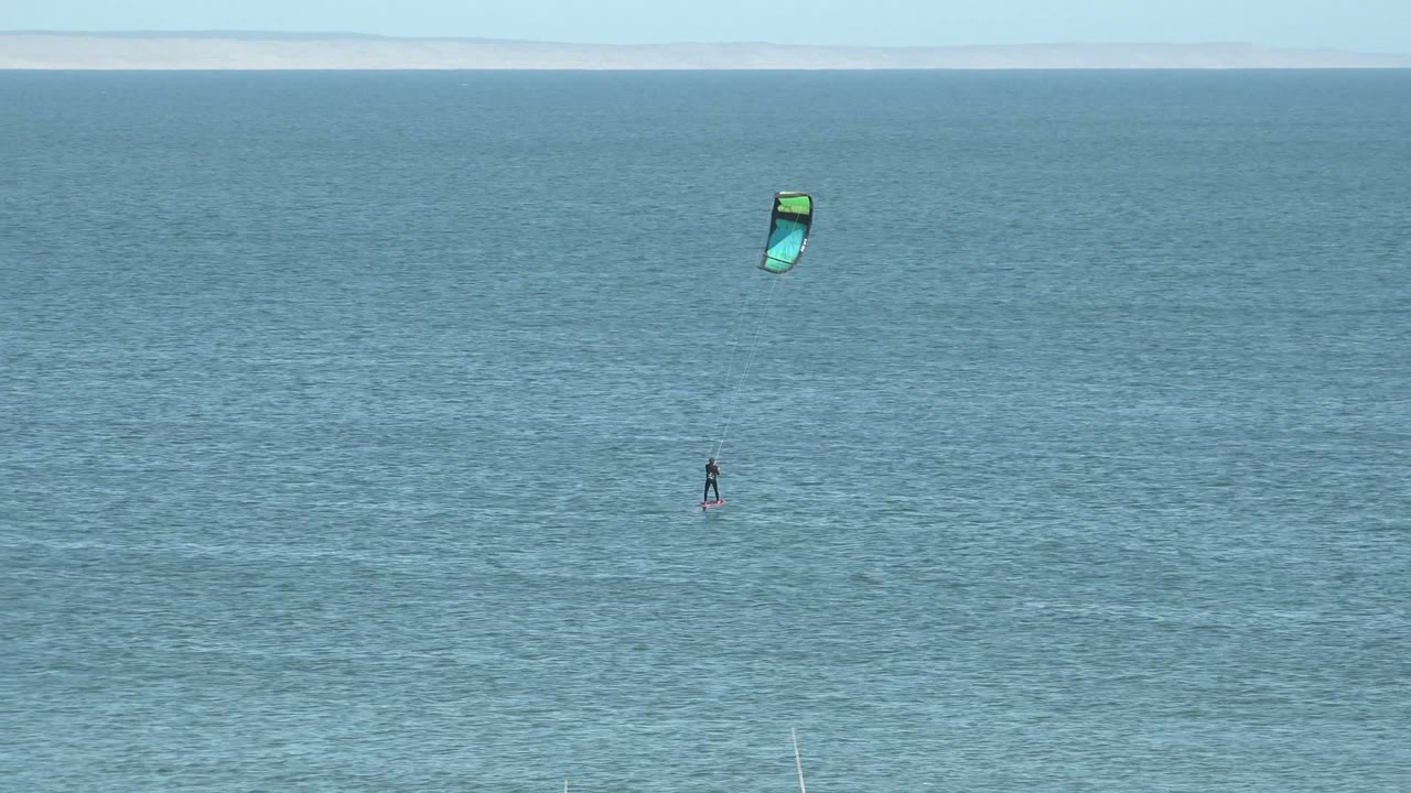 A single kite surfer remains distant on calm blue ocean in San Juanico, with the green and blue kite slightly lower in the sky than in the previous moment.