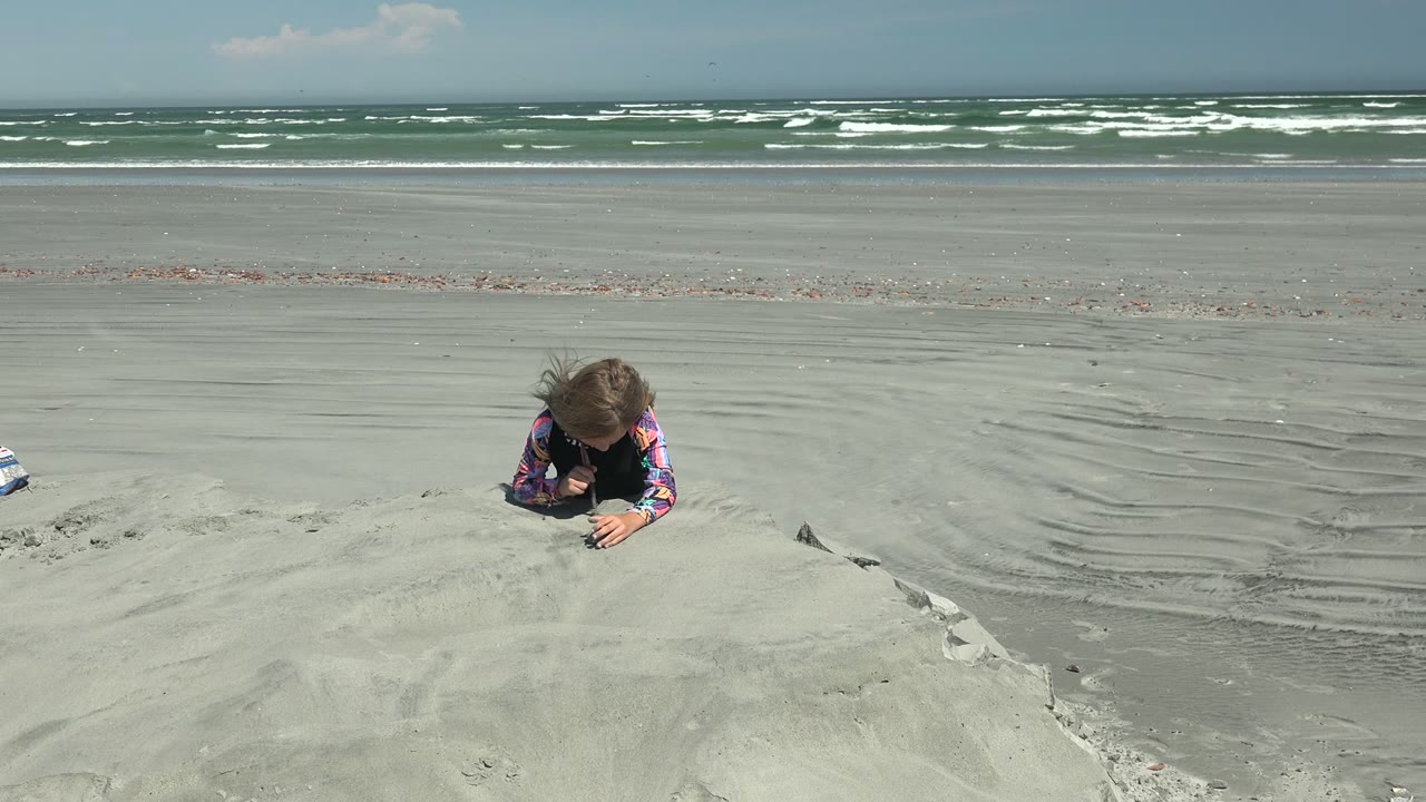 At 0:00 in La Bocana, Mexico, a child leans over a foreground sand mound while white surf breaks across the distant shoreline.