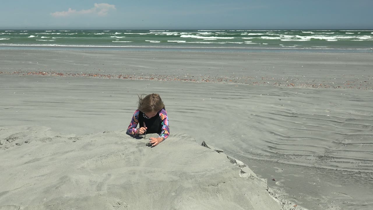At 0:02 in La Bocana, Mexico, the child crouches over the mound as shallow sand ripples and distant breaking waves divide the frame.