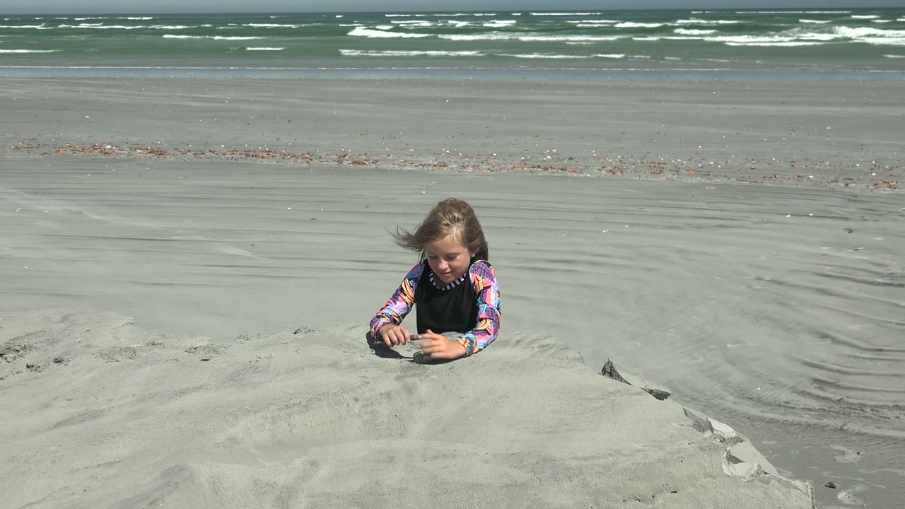 At 0:00 in La Bocana, Mexico, a child lies low behind a sand mound while waves break across the distant shoreline.