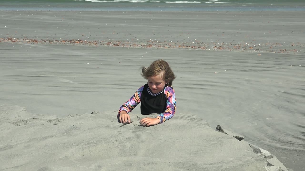 At 0:02 in La Bocana, Mexico, the child reaches forward across the sand mound, with pale beach flats and white wave bands in the background.