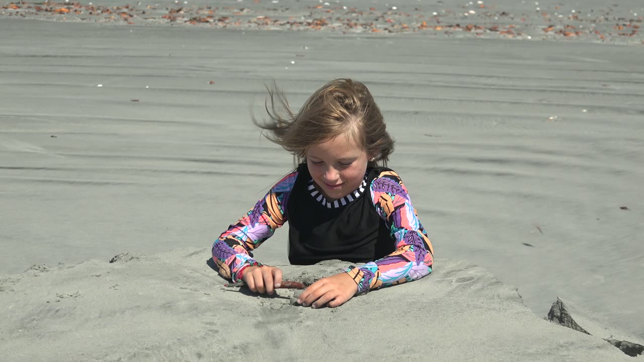 A child in a black vest and colorful long-sleeve shirt digs into pale sand on the beach at La Bocana, Mexico, with wind pushing hair to one side.