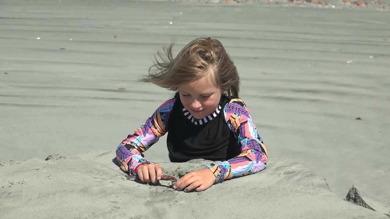 The child leans forward over a small sand mound in La Bocana, Mexico, keeping both hands low as the bright sandy shore fills the background.