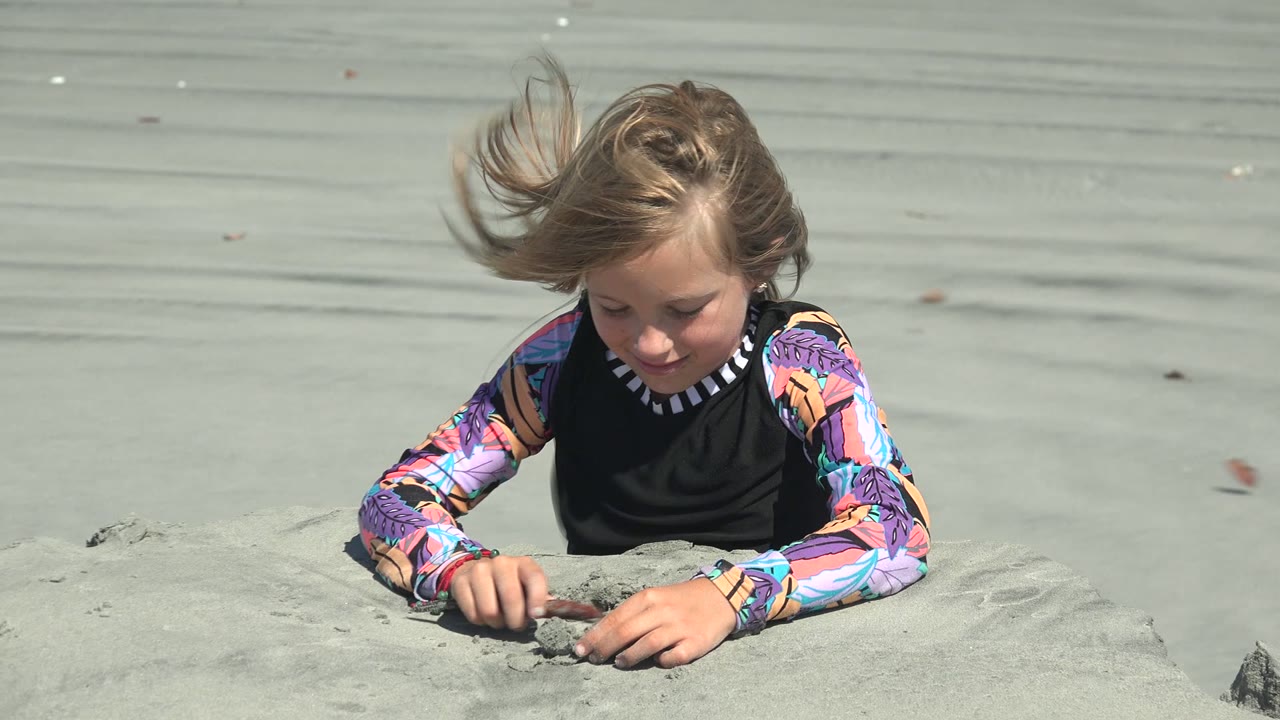 A gust lifts the child's hair while they continue pressing and scraping sand with both hands on the La Bocana beach.
