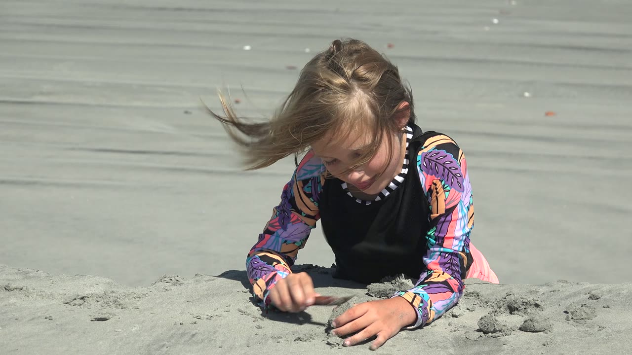 At 0:00 in La Bocana, Mexico, a child in a colorful long-sleeve shirt leans low over pale sand and presses a small mound with one hand.