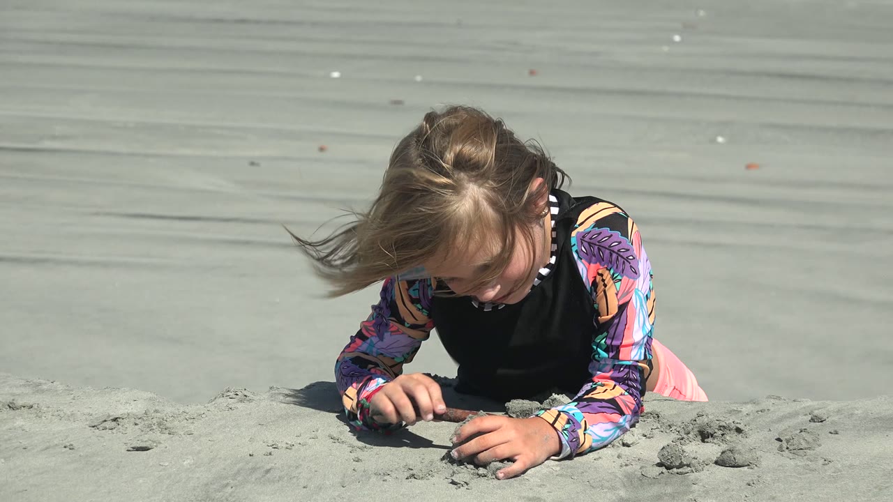 At 0:02 in La Bocana, Mexico, wind pushes the child's hair sideways as they bend over a small sand shape near the foreground edge.