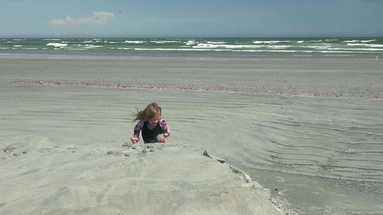 At La Bocana, Mexico, a child kneels low behind a sand mound near the left side of frame as white-capped waves line the distant shore.