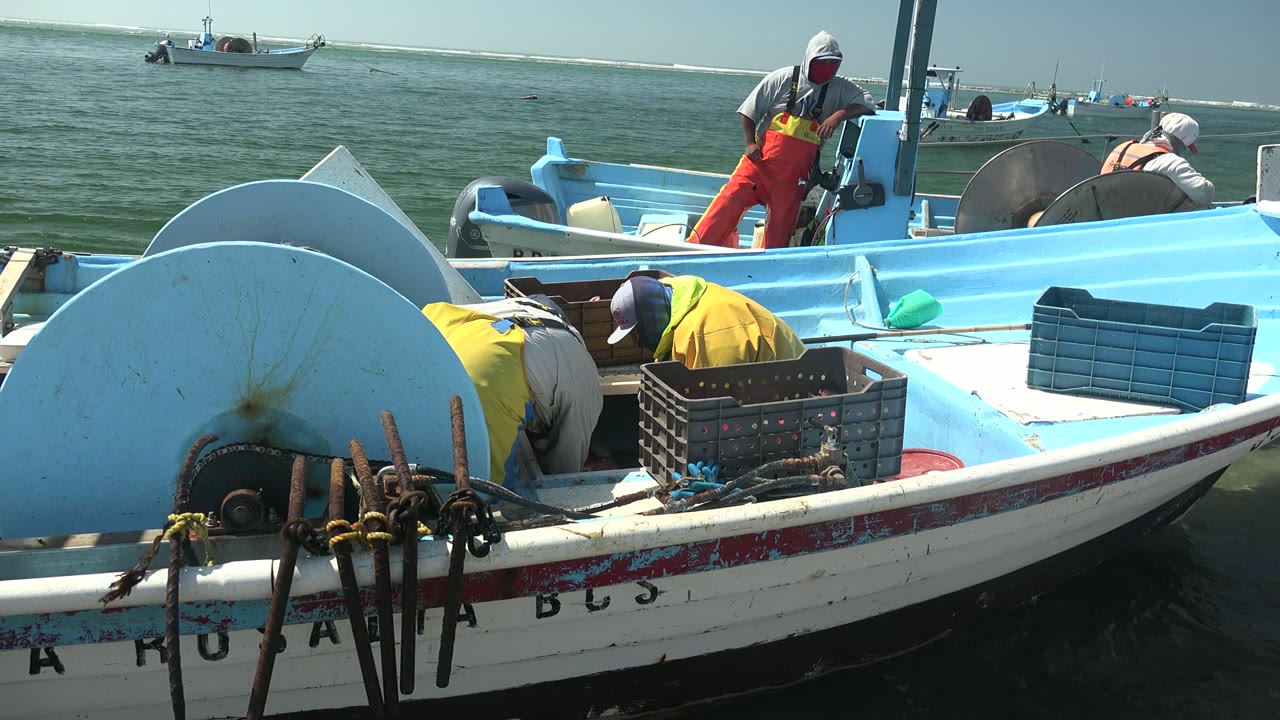 At 0:02 in La Bocana, the foreground shows the boat side and bundled rusted gear while crew members continue working around crates on the light blue deck.