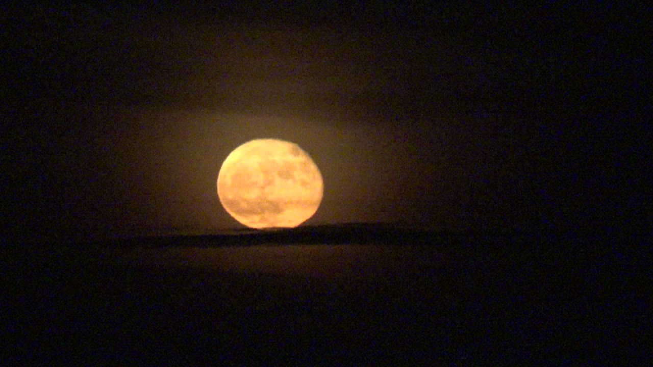 Orange full moon sits low above a dark horizon in La Bocana, Baja California Sur, with a faint glow in the night sky and no visible people.