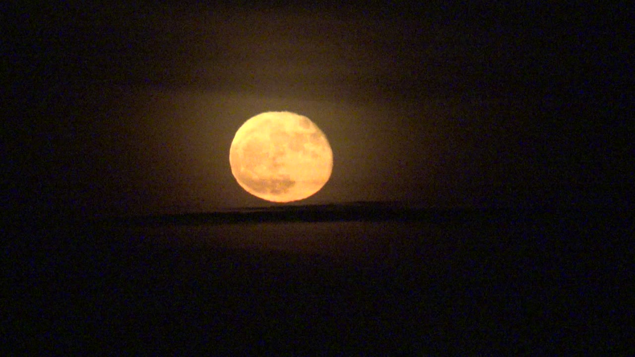 At 0:00 in La Bocana, Baja California Sur, Mexico, an orange full moon glows above a dark horizontal cloud band in a black night sky, with no visible people.