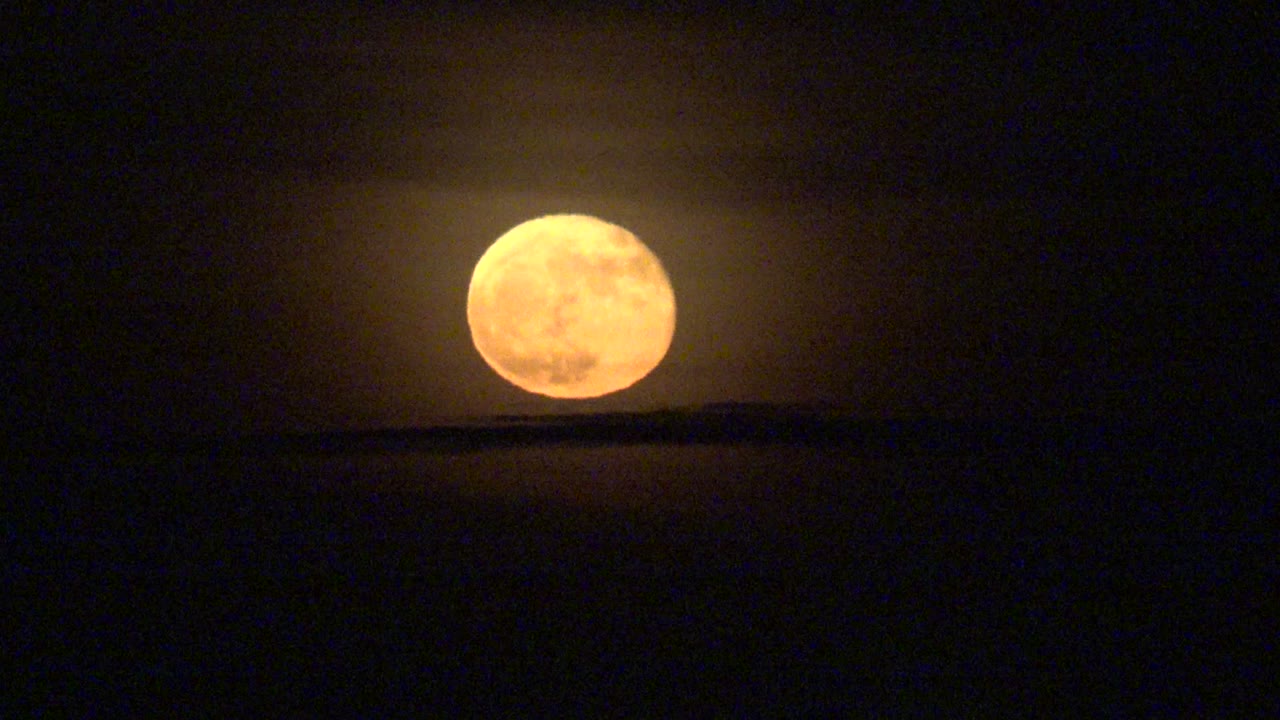 At 0:00, an orange full moon sits just above a low dark horizon in La Bocana, Baja California Sur, with no visible people.