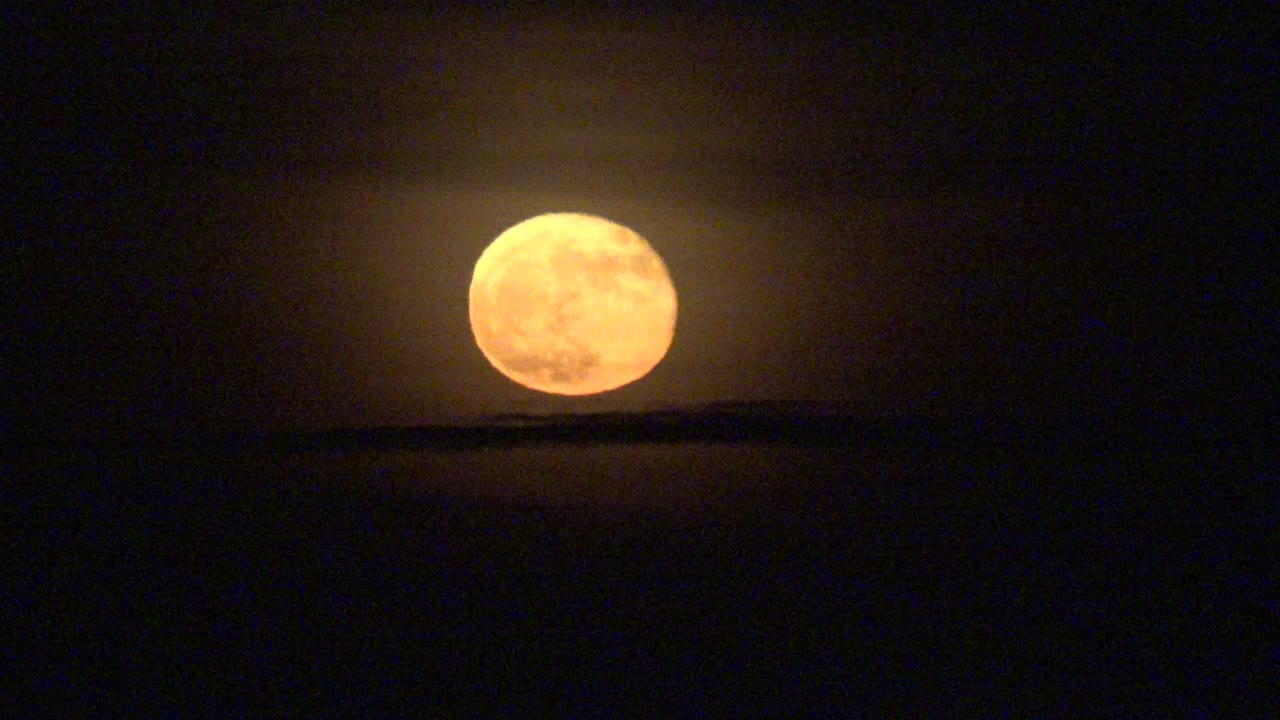 At 0:02, the orange full moon glows above a flat, dark horizon in La Bocana, Baja California Sur, surrounded by faint haze and no visible people.