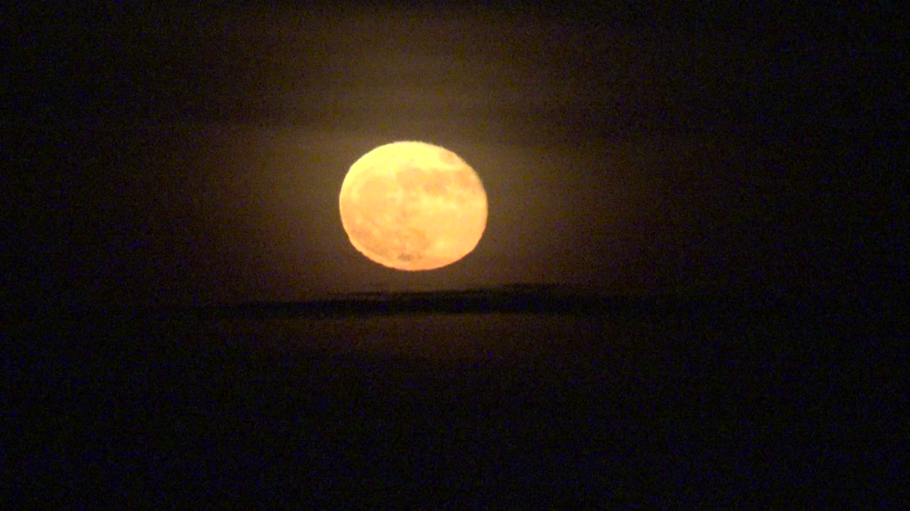 Orange full moon hangs low over a dark band in the night sky at La Bocana, Baja California Sur, with no visible people.