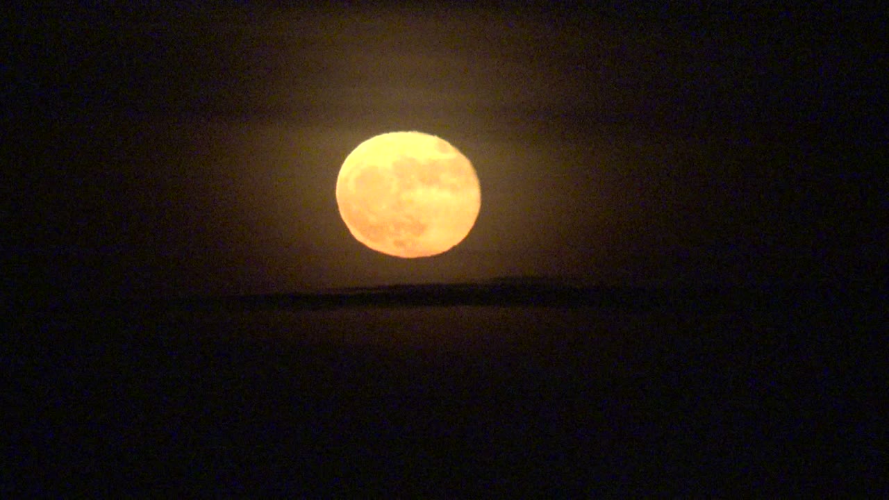 At 0:02, the orange full moon appears slightly right of the earlier framing above the dark La Bocana horizon, with thin haze crossing the moonlit sky and no visible people.