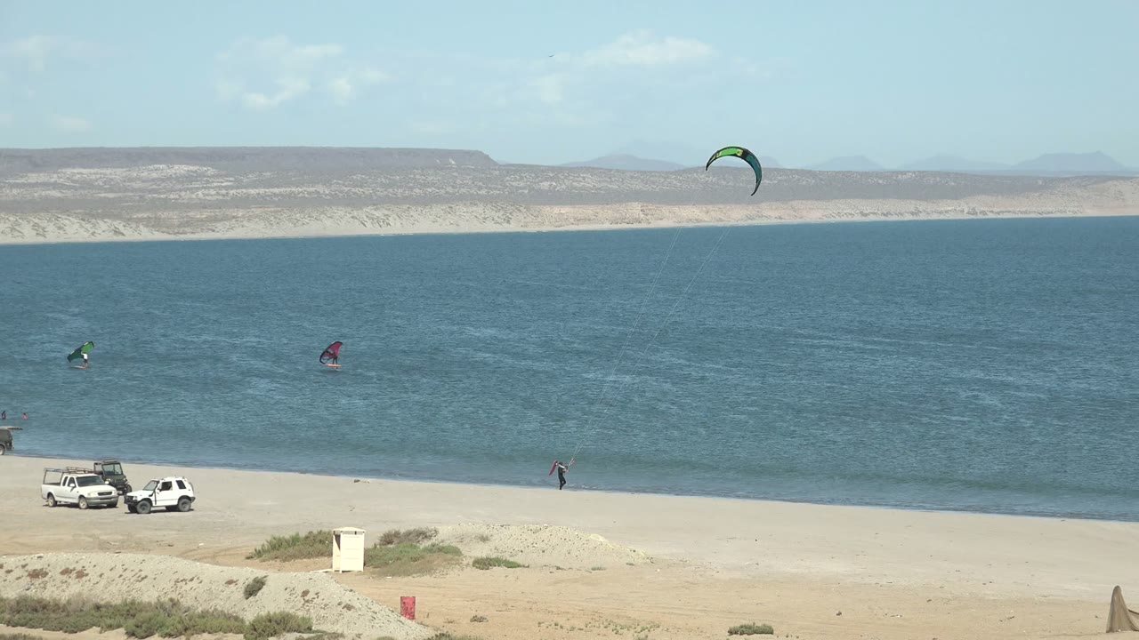 At 0:00 in San Juanico, Baja California Sur, a wide beach frame shows green and red kite surfing kites over blue water, white vehicles on the sand, small people near the shore, and distant hills.