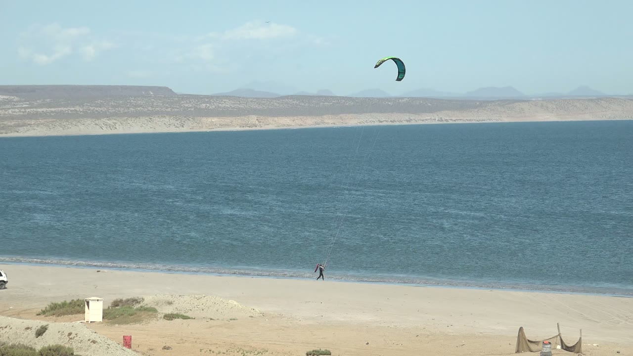 A green kite arcs high over a sandy San Juanico beach, with a small person near the shoreline, blue water, distant arid hills, and low beach items at the foreground edge.
