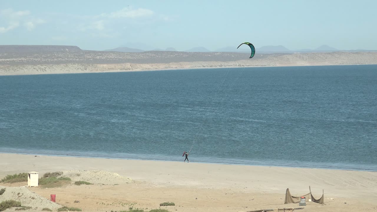 A wide San Juanico beach frame places the small rider near the waterline, the green kite near the upper sky, and calm blue ocean before muted arid hills.