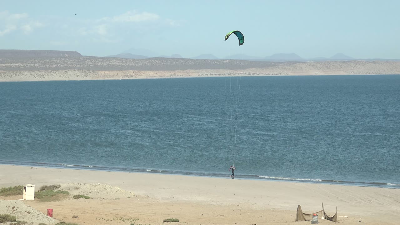 Wide San Juanico beach frame with a distant person near the shoreline, a green kite above blue water, pale sand in front, and low mountains across the horizon.