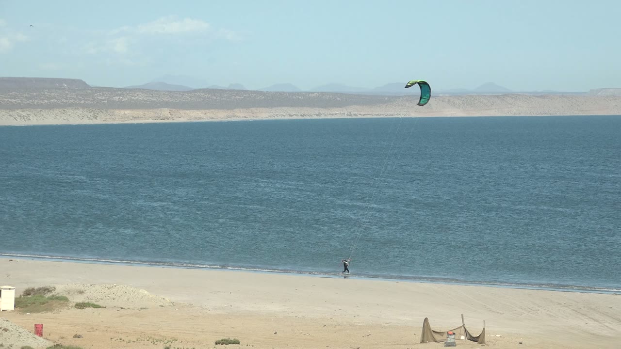 Wide San Juanico beach frame showing the rider farther right along the shoreline, a green kite near the upper right sky, blue water, pale sand, and low mountains in back.