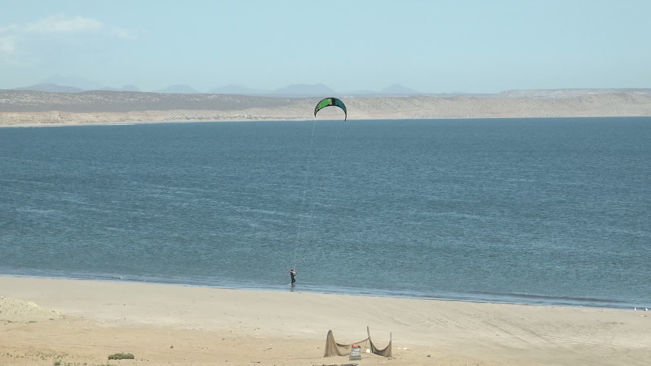 A distant kitesurfer stands near the waterline in San Juanico, Baja California Sur, with a dark and green kite high over calm blue water and pale sand in the foreground.