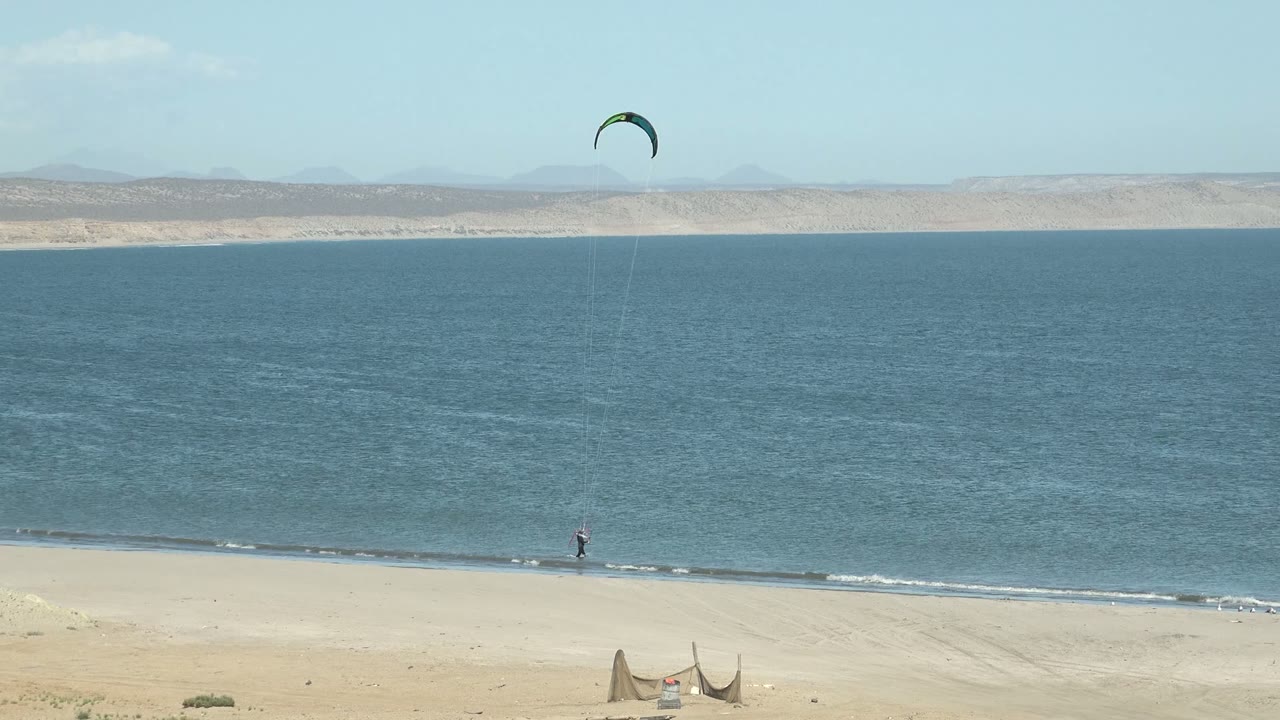 A tiny kitesurfer moves along the San Juanico waterline while the kite sits high toward the right side of the frame above calm ocean and a pale desert coastline.