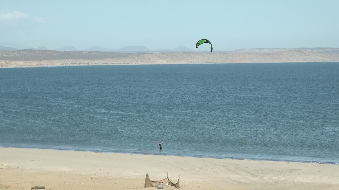 At 0:00 in San Juanico, Baja California Sur, a green and dark kite hangs over blue water while a very distant kite surfer stands near the shoreline, with pale sand below and low hills across the horizon.