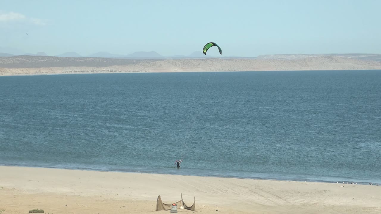 At 0:01 in San Juanico, Baja California Sur, the kite sits high over the water and the distant surfer remains near the waterline as small waves meet the sandy beach.