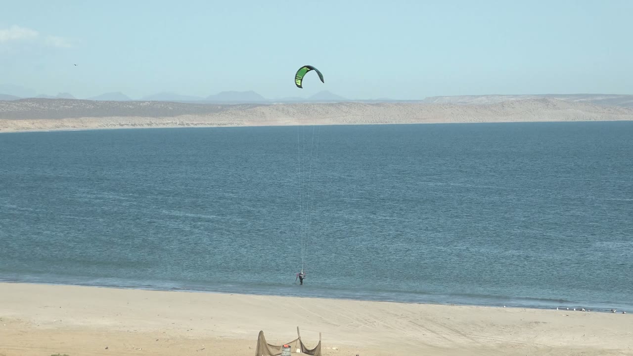 At 0:02 in San Juanico, Baja California Sur, the kite drifts near the upper right of the frame while the tiny surfer is close to the breaking waves below.