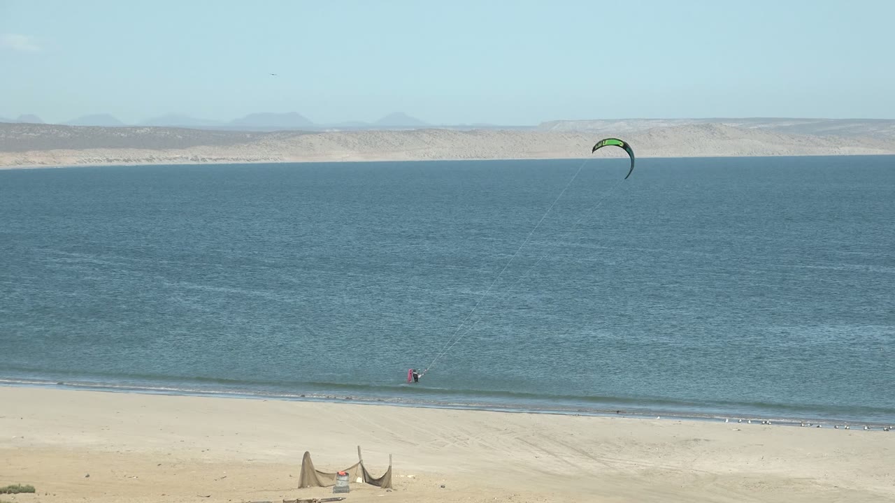 Green kite over blue water near a sandy San Juanico beach, with distant mountains on the horizon and no recognizable people visible.