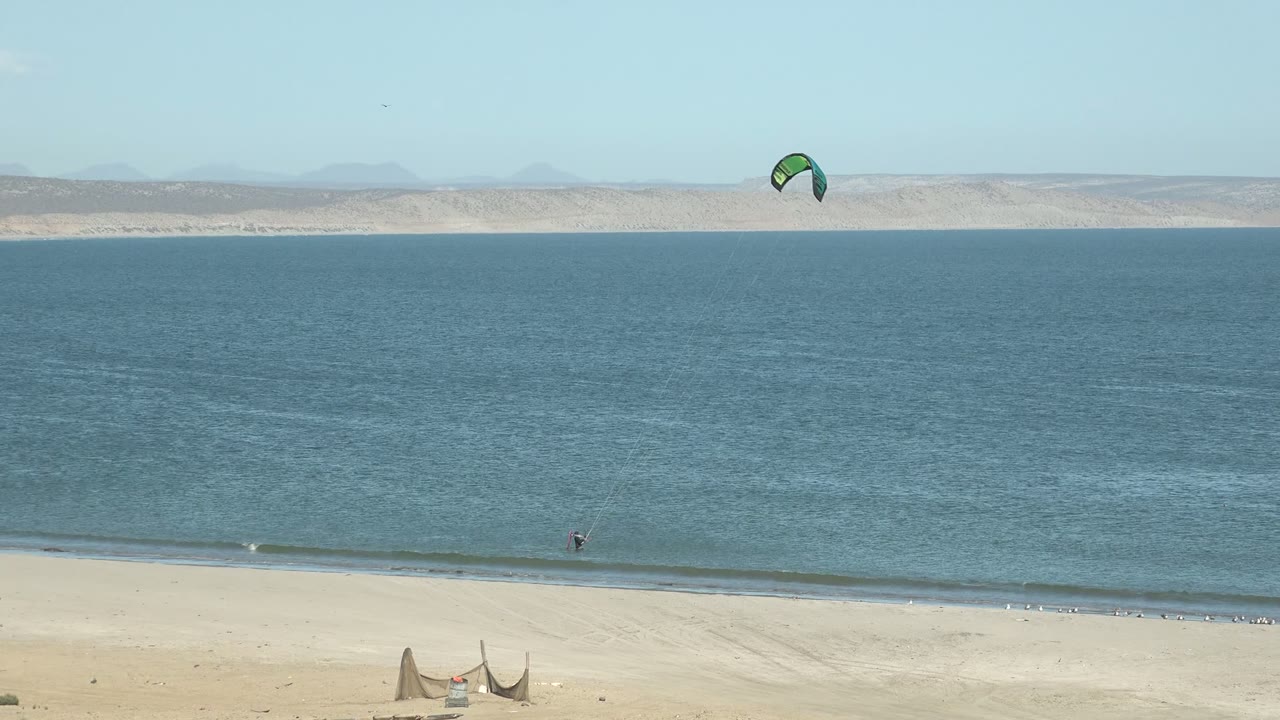 A kite rides high above the San Juanico shoreline while a tiny kite surfer appears near the surf line, with pale sand and low waves below.