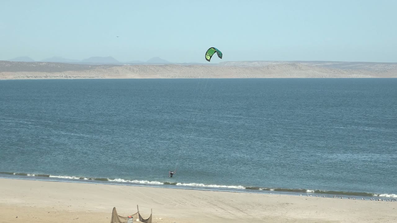 The kite sits near the upper right of the San Juanico frame above open blue water, with a narrow line of breaking waves on the beach.