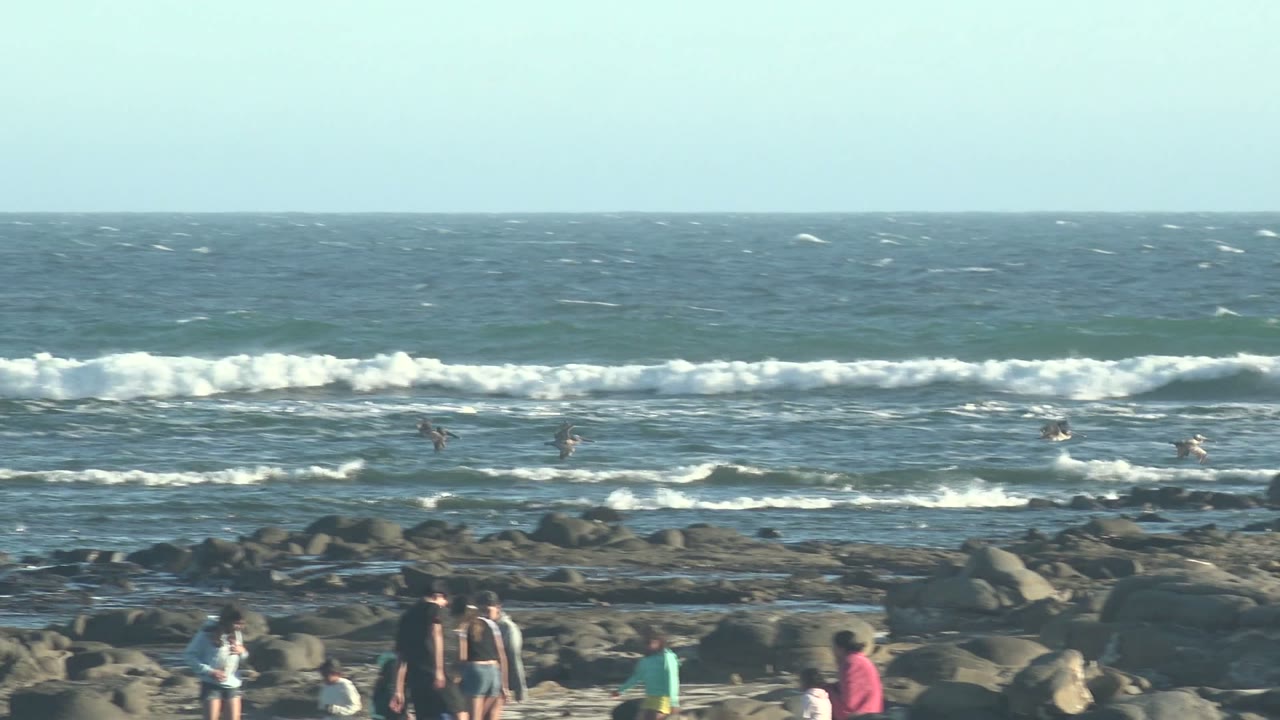 Right-side La Bocana beach view with several people on dark rocks and multiple white wave bands rolling toward shore.