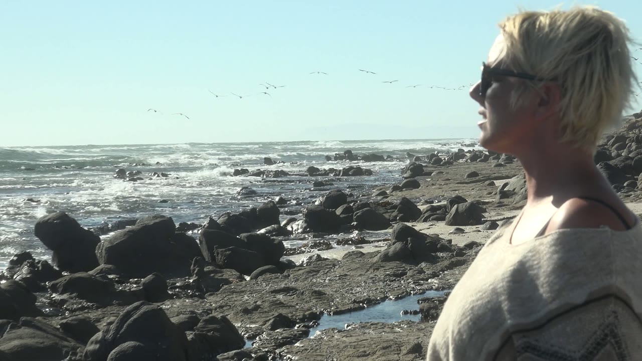 At La Bocana, Mexico, a blonde woman in sunglasses stands in left profile near dark shoreline rocks while waves break across the background.