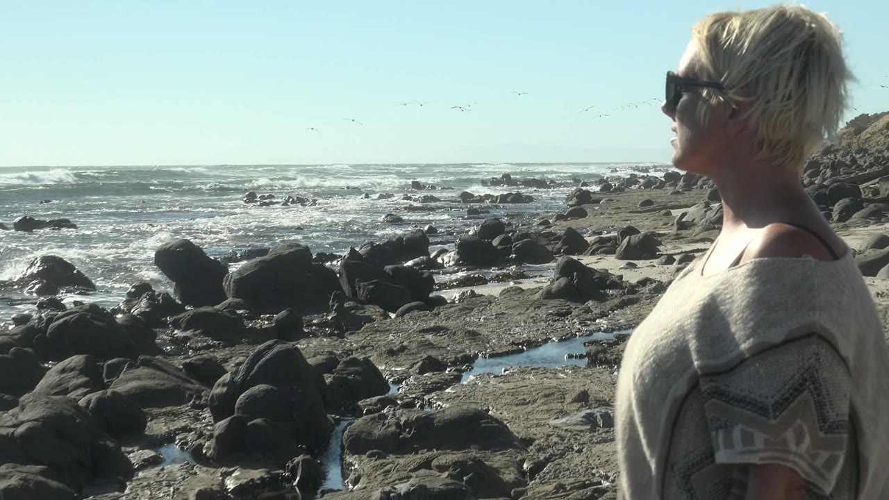 Woman facing the ocean with rocks and surf behind her.