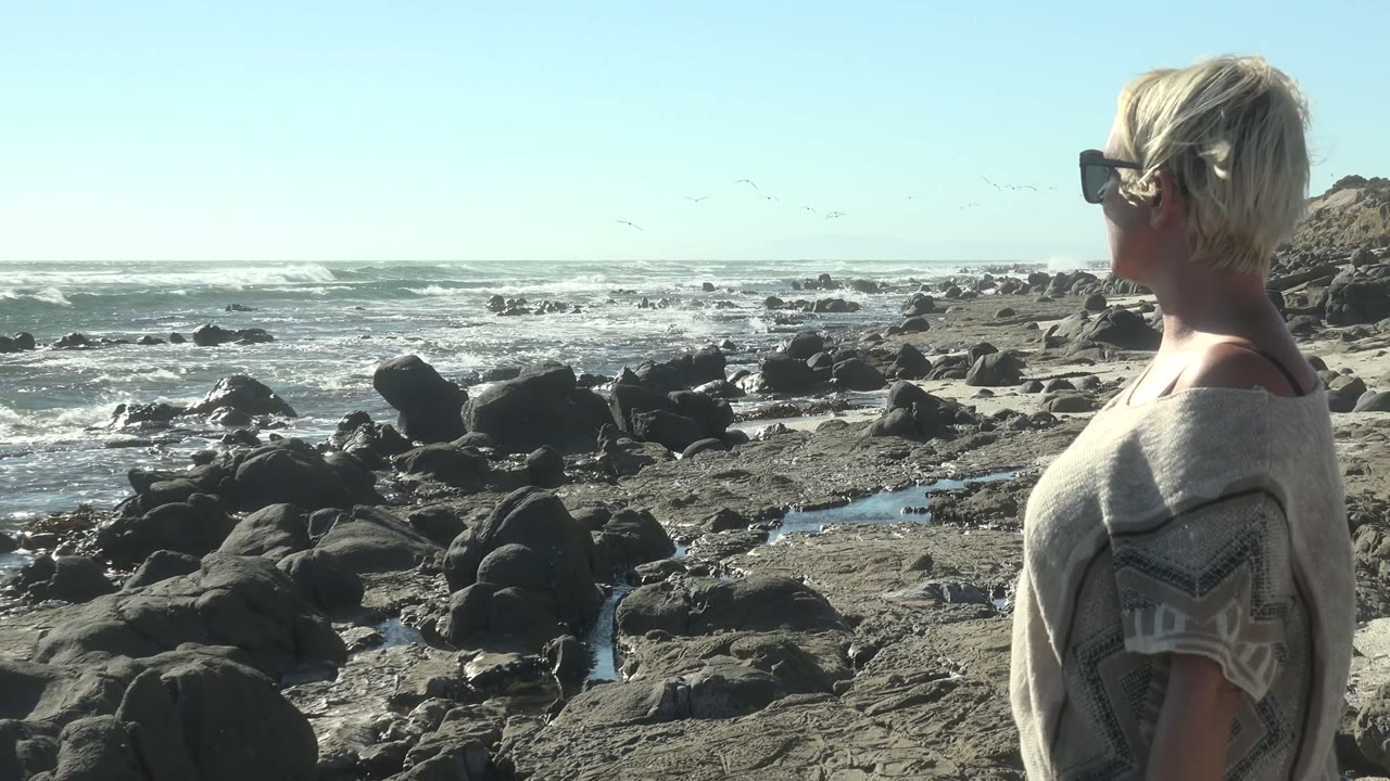 At La Bocana, Mexico, the woman is framed farther to the right with more rocky beach and shallow pools visible between her and the foamy ocean.