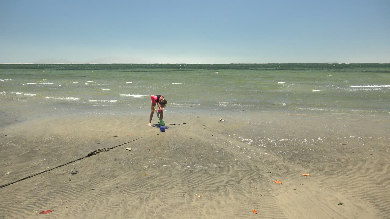 At 0:00 in San Carlos, Baja California Sur, a child in a red top bends over a blue bucket on wet sand while small waves roll across the beach behind them.