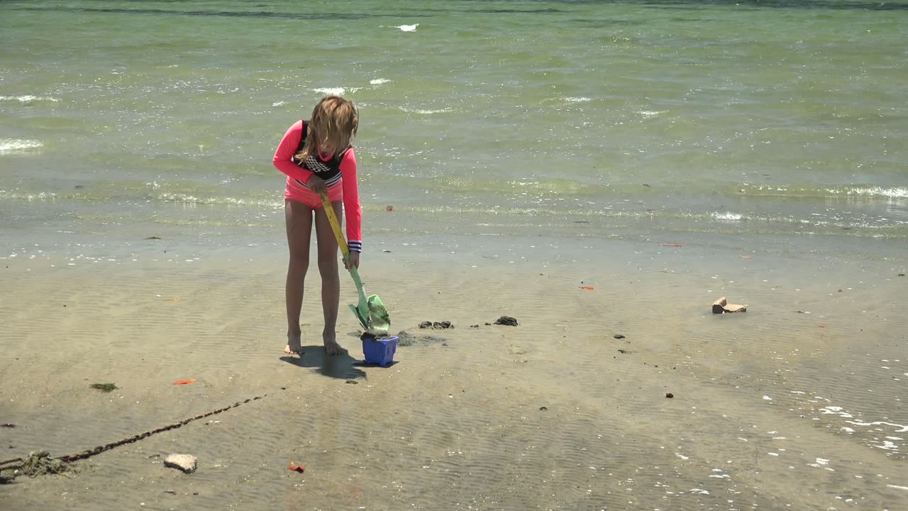 At 0:00 in San Carlos, Baja California Sur, a child in a pink and black swimsuit stands over wet sand with a green shovel and blue bucket near small shoreline waves.