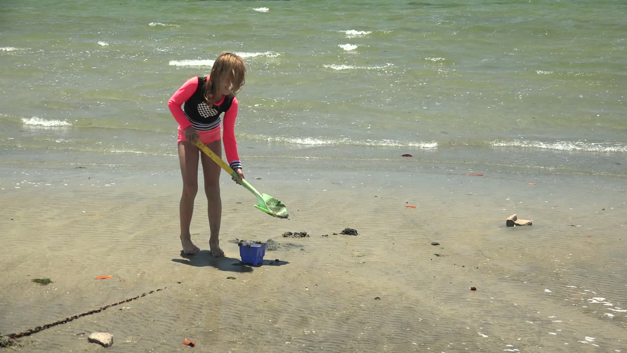 Child holds a green shovel over wet beach sand beside a blue bucket.