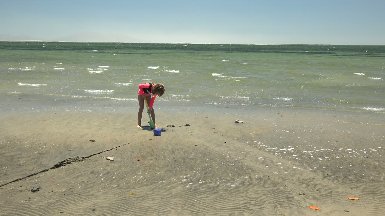 At the start of the San Carlos, Mexico clip, a child in a red shirt bends low over wet sand beside a blue bucket while small ocean waves roll behind.