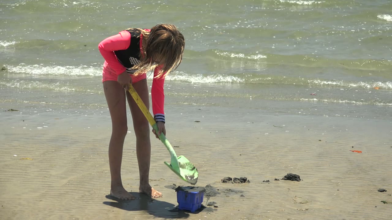Girl leans over a blue bucket with a green shovel on wet beach sand.