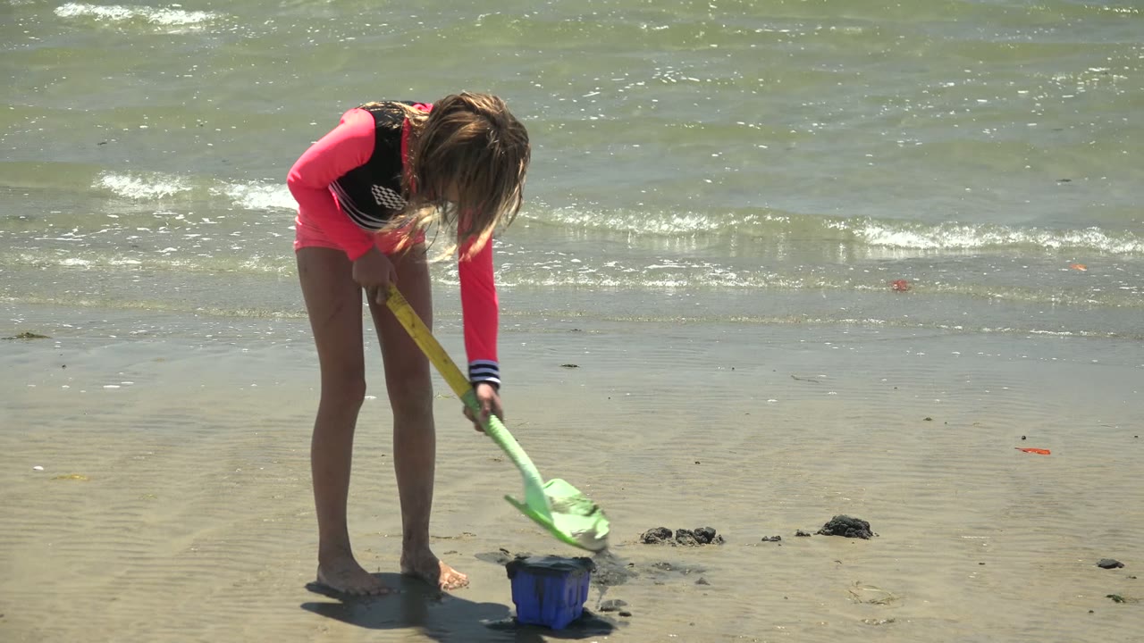 A young girl in bright pink beachwear lowers the green shovel toward the blue bucket on the wet San Carlos shoreline, with white ripples visible on the water behind her.