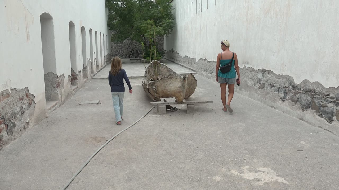 At 0:00 in Mulege, Mexico, a woman and child walk away from the camera toward a large wooden boat set between white courtyard walls.