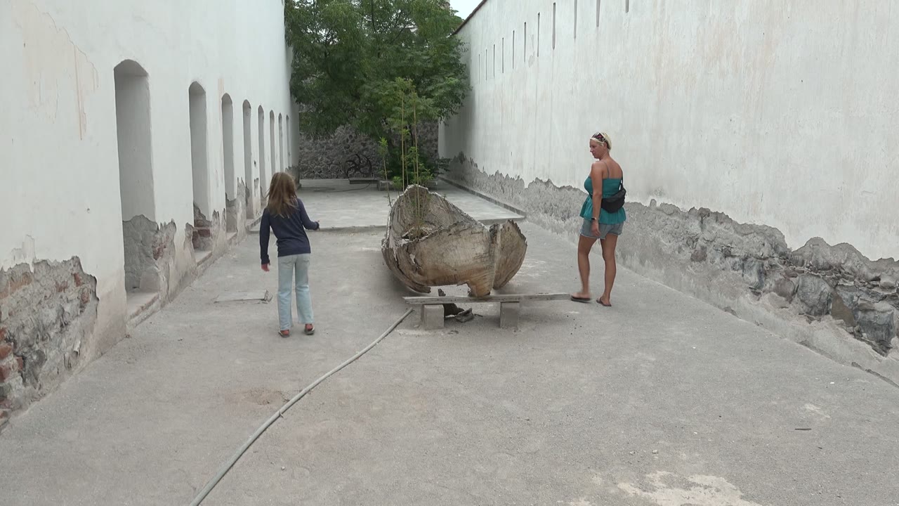 Child and woman pause near a large wooden boat beside white walls.