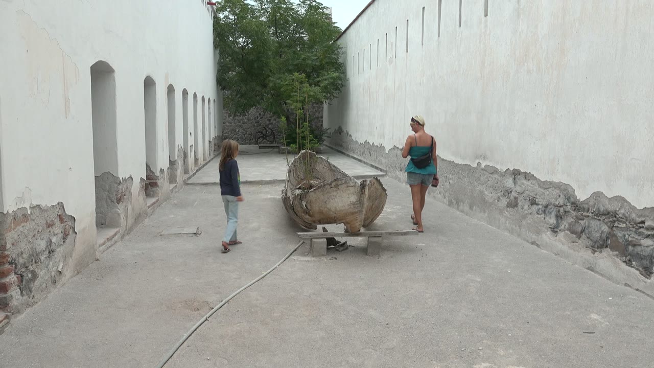 At 0:02 in Mulege, Mexico, the child stands left of the boat and the woman stands to the right, with the white walls forming a long corridor behind them.