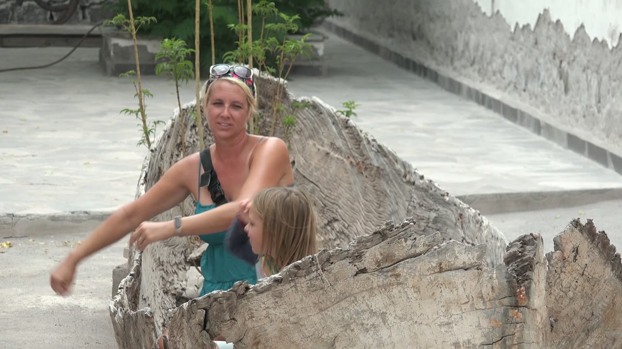 A woman in a teal top and a child sit within a large hollow tree trunk in a Mulege, Mexico courtyard as the woman extends one arm.