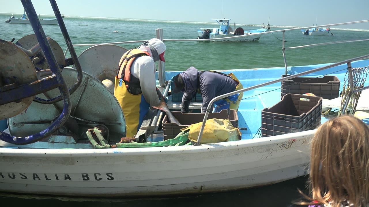 At La Bocana, one fisherman bends into the boat while the other leans over the deck, with a partial child head in the right foreground and small boats crossing the background water.