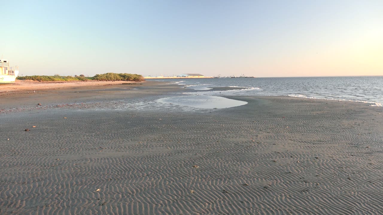 At 0:00 in San Carlos, Baja California Sur, rippled wet sand leads toward a shallow tide pool and small ocean waves, with part of a white boat still visible at the far left and no visible people.