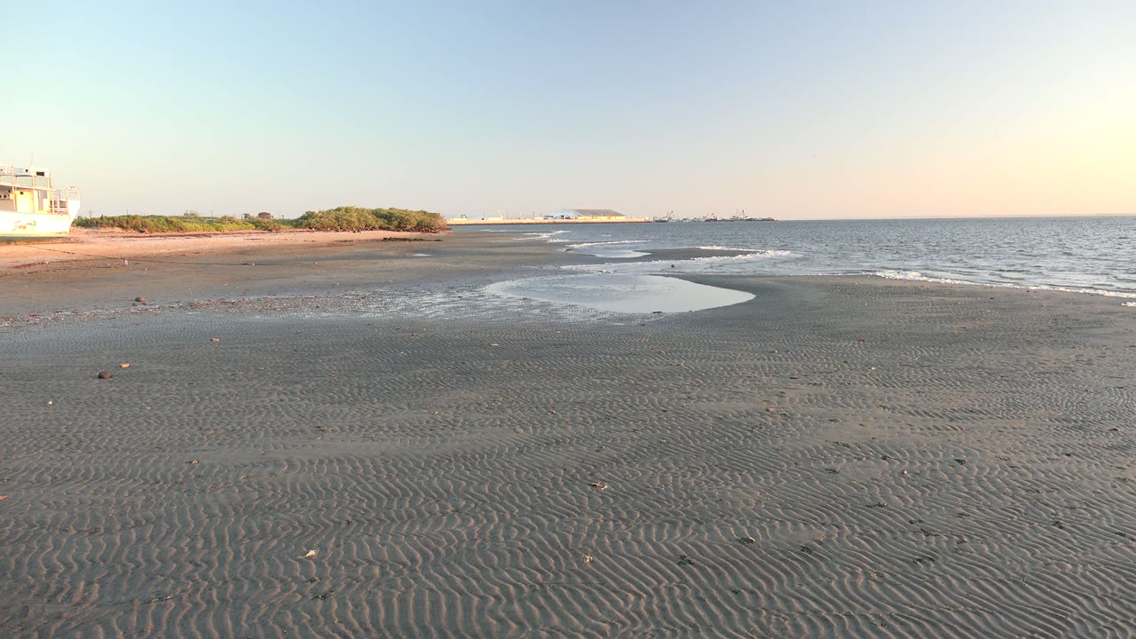 At 0:02 in San Carlos, Baja California Sur, the frame holds on empty rippled sand, a bright curved sheet of shallow water, small waves to the right, and low vegetation to the left with no visible people.