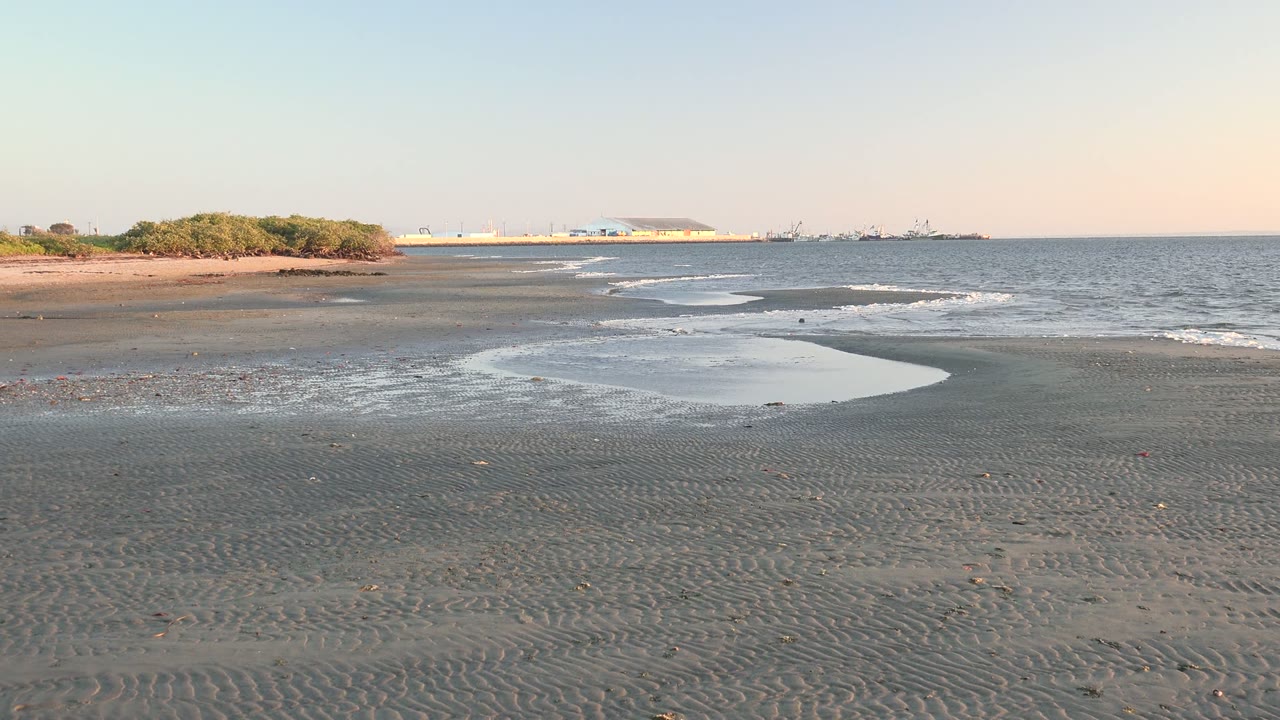Small waves lap across dark sand with a tide pool and distant buildings beyond.