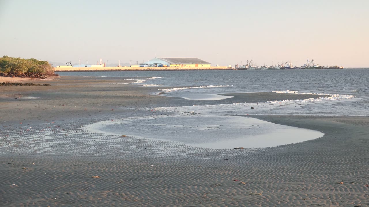 At 0:00 in San Carlos, Baja California Sur, dark rippled wet sand and shallow pools lead toward small waves, distant coastal buildings, boats, and no visible people.